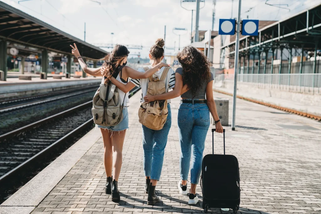 Trois jeunes voyageuses de dos, bras dessus bras dessous sur un quai de gare, portant des sacs à dos et une valise à roulettes, l'une d'elles faisant un signe de la main.