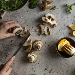 Vue de dessus de mains découpant des champignons de Paris sur un plan de travail, entourés d'épinards frais, de persil et de quartiers de citron.
