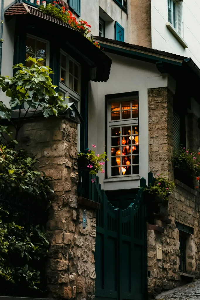 Façade d'une maison de charme accueillante, en pierre avec un portail vert foncé, des jardinières fleuries aux fenêtres et des lampions chaleureux éclairés à l'intérieur.